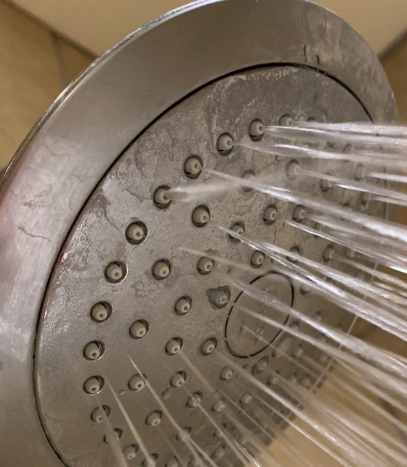 Shower head clogged from hard water minerals, with some jets not functioning and others spraying diagonally